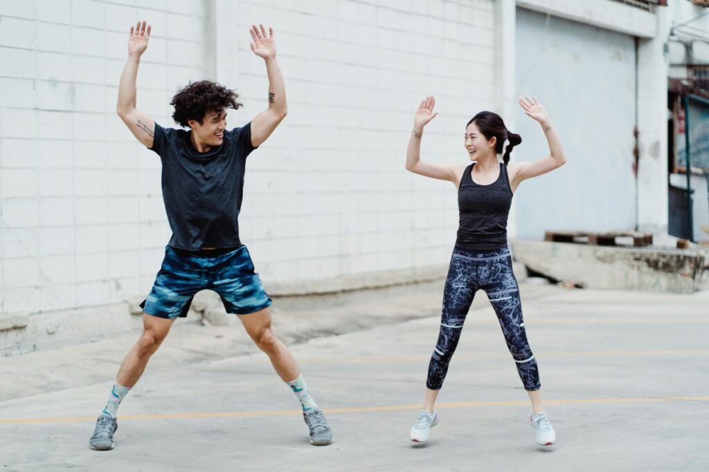 Boy and girl performing jumping jacks with arms raised and feet apart.